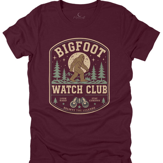 A black short-sleeve crewneck t-shirt featuring a vintage-style graphic print of Bigfoot in a tree forest under a full moon, with the text 'BIGFOOT WATCH CLUB' and 'BELIEVE THE LEGENDS'.