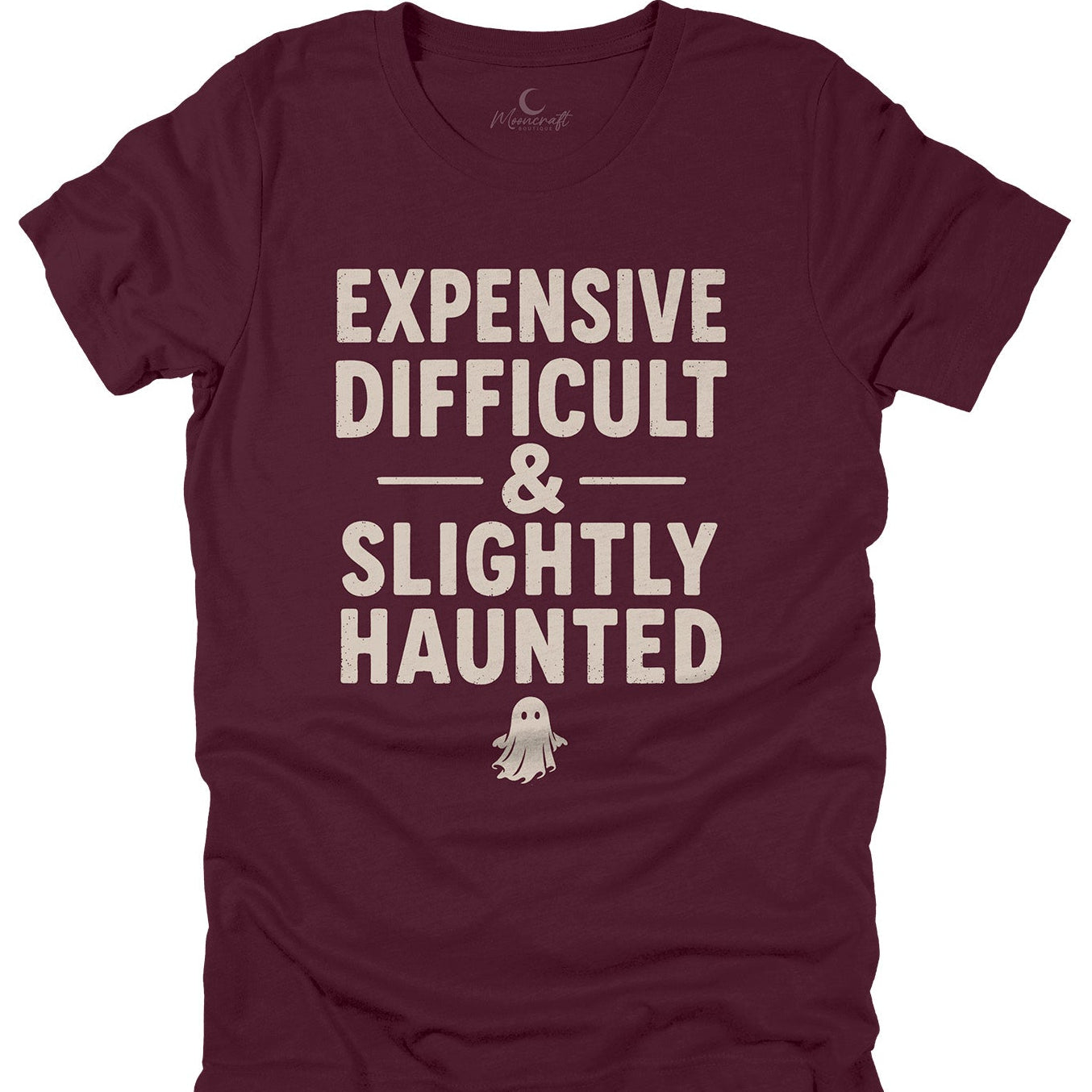 A maroon short-sleeve t-shirt with a cream-colored graphic text that reads 'EXPENSIVE DIFFICULT & SLIGHTLY HAUNTED' above a small ghost illustration, displayed flat against a white background.