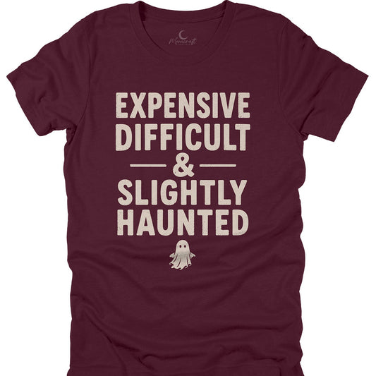 A maroon short-sleeve t-shirt with a cream-colored graphic text that reads 'EXPENSIVE DIFFICULT & SLIGHTLY HAUNTED' above a small ghost illustration, displayed flat against a white background.
