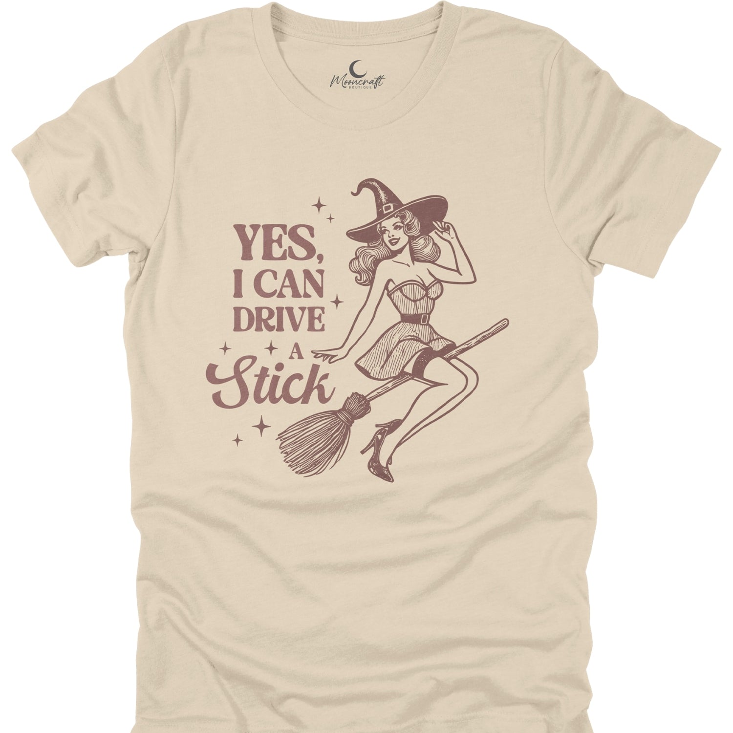 Beige t-shirt with a graphic of a witch on a broom and text 'Yes, I can drive a stick' on a white background.