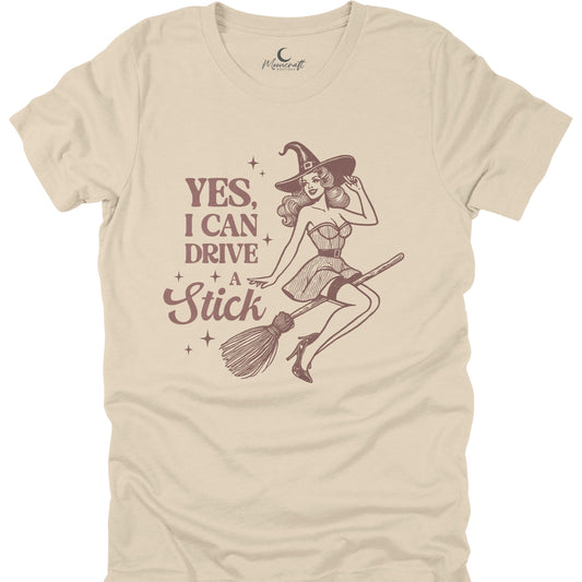 Beige t-shirt with a graphic of a witch on a broom and text 'Yes, I can drive a stick' on a white background.