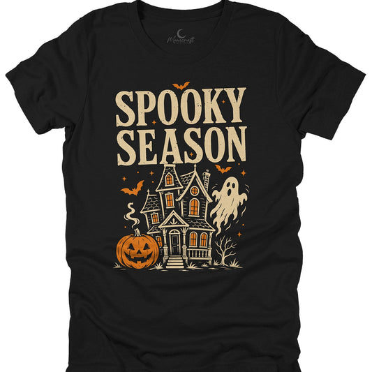 A black short-sleeve t-shirt with a 'Spooky Season' Halloween graphic, featuring a haunted house, jack-o'-lantern, ghost, and bats in beige and orange, displayed on a solid white background.
