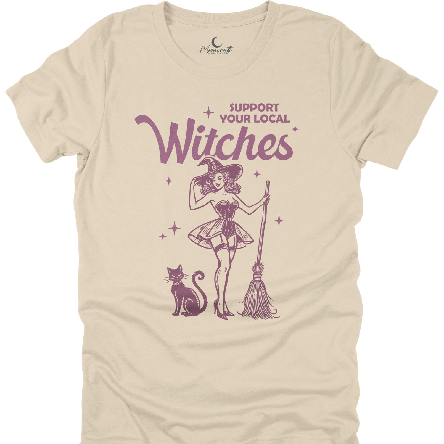 Beige t-shirt with a graphic of a witch, cat, and broomstick, and text 'Support Your Local Witches' on a white background.