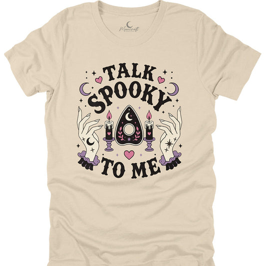A beige short-sleeve t-shirt with a gothic 'Talk Spooky To Me' graphic on the front, featuring a central apothecary triangle, candles, and witchy hands, displayed on a clean white background.